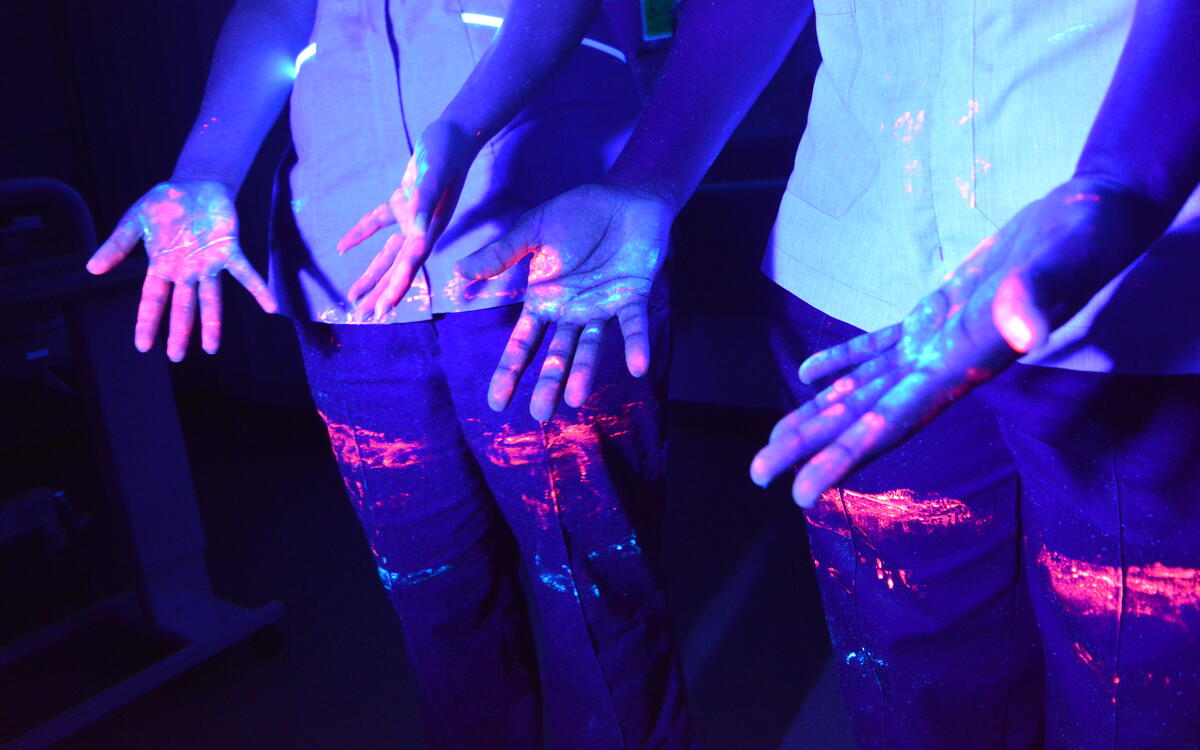 Hands glowing with different coloured mock-pathogens , lit under ultra-violet light.